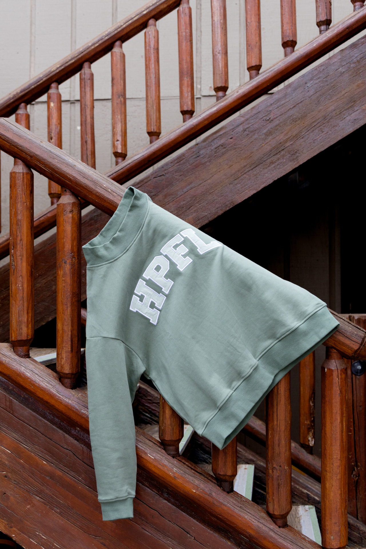 Sage green HPFL crewneck sweatshirt with white applique lettering hanging on a wooden staircase railing
