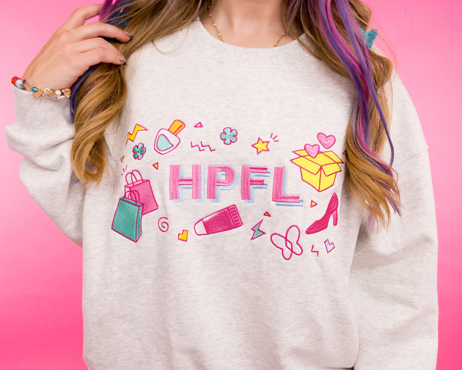 Close-up of a heather grey HPFL crewneck with colorful embroidered icons on a pink backdrop